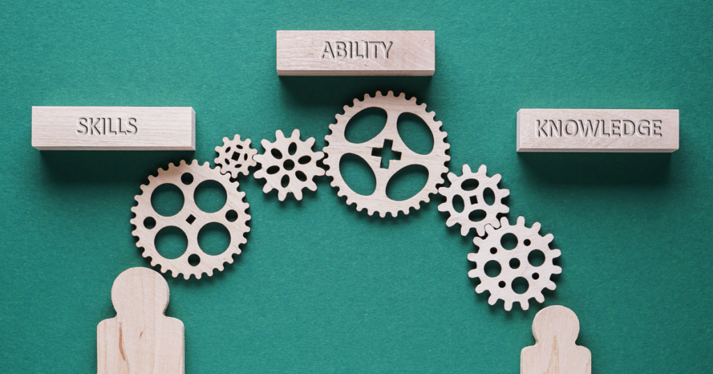 Photo of wooden figures underneath wooden gears with blocks named skills, ability, knowledge over them