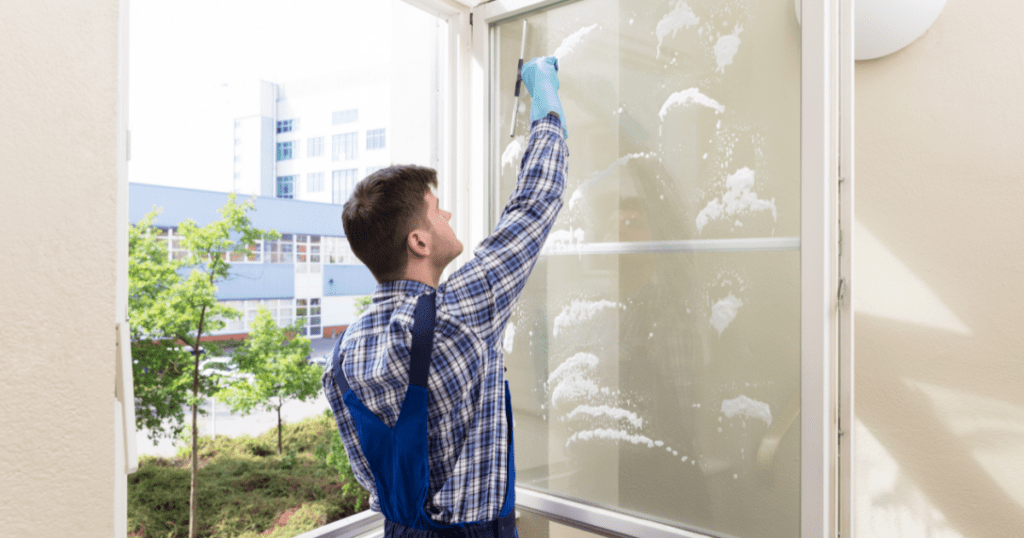 mancleaningwindows | Pathways Career College a man in blue overalls uses squeegee to clean office windows, overlooking green trees and buildings