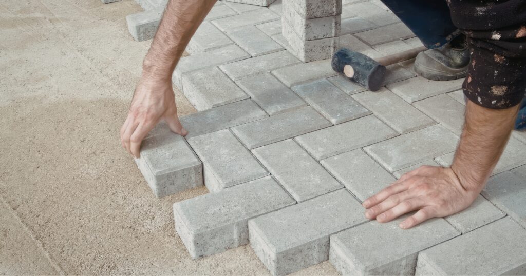 Bricklaying ‣ Pathways Career College Close-up of hands placing gray concrete pavers during patio construction on sandy ground.